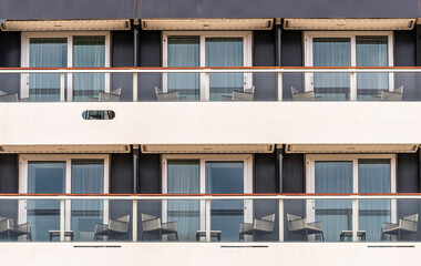 side of a cruise ship with balconies and deck chairs. Cabin windows with tables and seats on the...