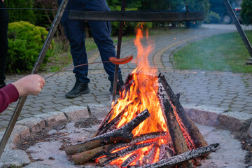 Sausage roasting over open campfire in backyard setting, people enjoying outdoor cooking with...