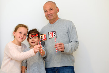 Happy grandfather with two granddaughters holding a decorative LOVE sign indoors. Cheerful family moment, warm bonding and joyful smiles, perfect for themes of love, celebration and togetherness.