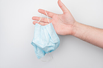 A close-up photograph of a hand holding a blue face mask with elastic bands The background is light...