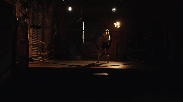 Authentic fight club, woman fighter training alone with punching bag in box ring . Cinematic slow motion shot in gym, beautiful slender lady striking by hands, practicing punches, preparing to fight