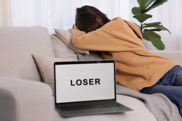 Cyber bullying. Upset teenage girl and laptop with text You Loser on sofa at home, selective focus