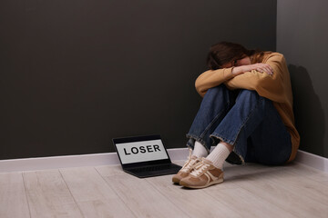 Cyber bullying. Upset teenage girl and laptop with text You Loser on floor indoors. Space for text