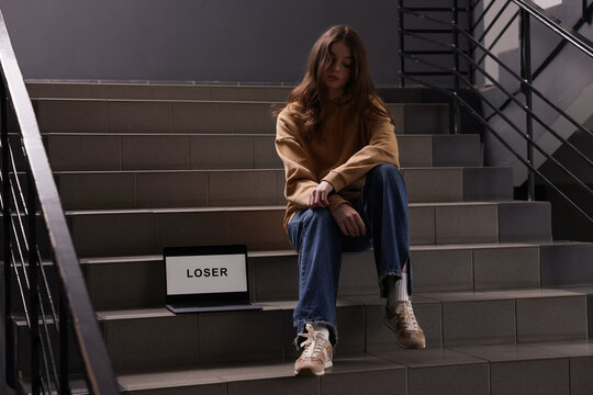 Cyber bullying. Upset teenage girl and laptop with text You Loser on stairs indoors