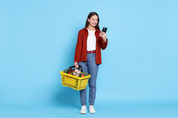 Smiling woman with basket full of products and smartphone on light blue background. Online shopping