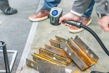 Person using a vacuum cleaner with a laser anti-corrosion tool in an industrial workshop Grey floor, dark metal components scattered on the floor, muted color palette with blue shirt as contrast