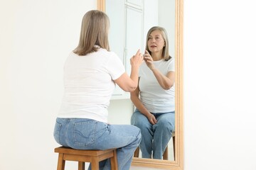 Beautiful senior woman looking at herself in mirror indoors