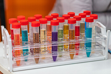 Collection of colorful glass test tubes arranged in a clear plastic holder, with diverse liquids ranging from blues, greens, yellows, reds to purples The setting appears indoor, lit artificially wit