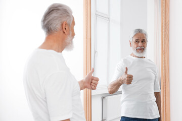 Handsome senior man showing thumbs up mirror indoors