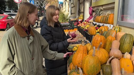 Two women are examining pumpkins at an outdoor market stall.  Autumn harvest abundance of various gourds, apples and grapes are visible. A vibrant autumn scene. - Powered by Adobe