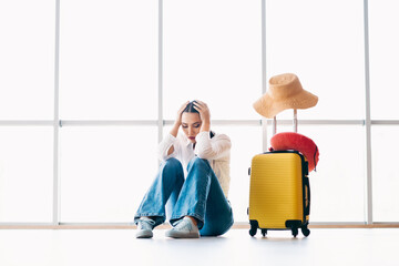 Sad woman sitting near suitcase in airport terminal during uncertain travel situation with casual...