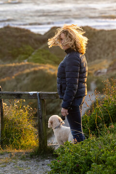 Hirtshals, Denmark A woman with blond hair blowing in the wind and a Bichon Poo dog overlloking the dunes of the North Sea.