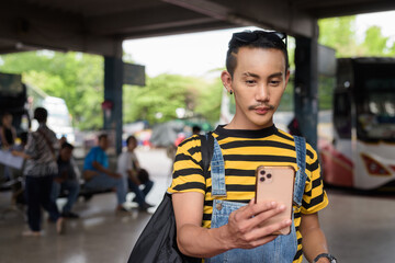 Portrait of nonbinary genderfluid young Asian gay tourist man in bus terminal using phone
