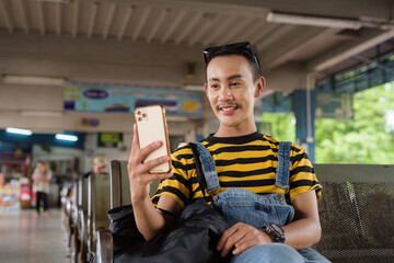 Portrait of nonbinary genderfluid young Asian gay tourist man in bus terminal using phone