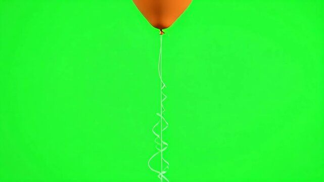A single orange balloon floating in front of a bright green screen in a studio setting view