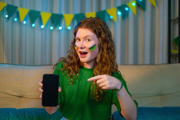 Red-haired woman with green face paint pointing to a blank smartphone screen, ready for mobile app...
