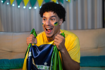 Male soccer fan with Brazilian face paint and yellow shirt watching intensely while clutching a...