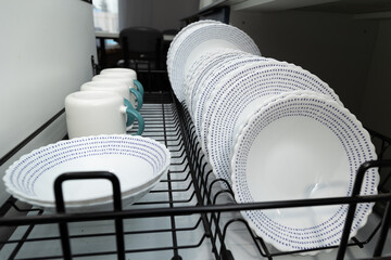 Clean, white plates and bowls with blue accents are drying on a metal rack next to mugs in a cozy...
