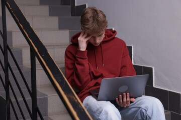 Cyber bullying. Upset teenage boy with laptop on stairs indoors