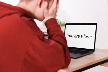 Cyber bullying. Upset teenage boy and laptop with text You Loser at desk indoors, closeup