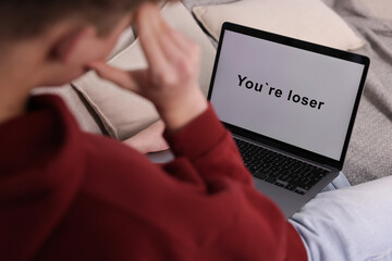 Cyber bullying. Upset teenage boy looking at laptop with text You Loser on sofa at home, closeup