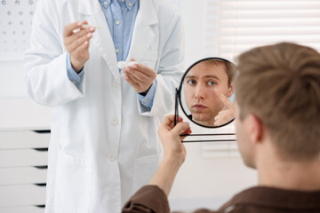 Fototapeta premium Young man putting on contact lenses near mirror in ophthalmologist office, selective focus