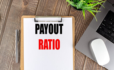 Clipboard with text payout ratio on wooden desk next to laptop, pen, mouse and plant