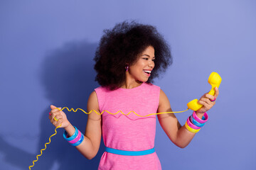 Cheerful young woman in pink dress with afro hairstyle holding a yellow phone in hand against a...