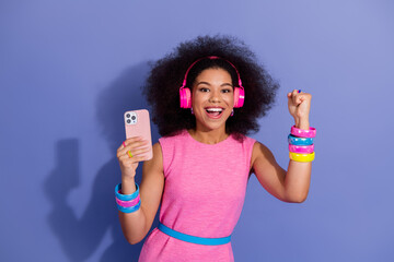 Excited young woman in a bright retro outfit holding a smartphone and posing on a vibrant violet...