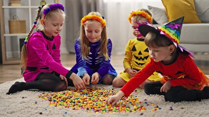 Children in colorful Halloween costumes collect candies from the carpet at home