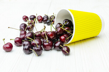 Fresh cherries poured from yellow cup on white wooden background