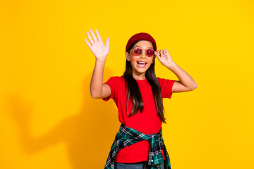 Cheerful young girl in red t-shirt and sunglasses waving hand on bright yellow background, showing...