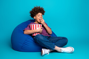 Young boy relaxing on a bean bag chair holding a popcorn bucket against a vibrant blue background