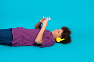Smiling teen boy lying on blue background, yellow headphones and phone in hand, casual and cheerful...