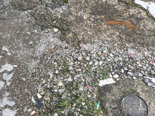 Top view of cracked asphalt surface with loose gravel, small plants, and urban litter.
