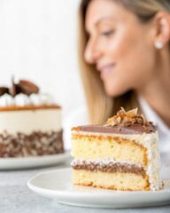 A close-up of a slice of layered cake with chocolate frosting and nuts, with a smiling woman blurred in the background.