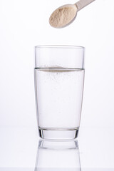 Collagen powder is poured with a measuring spoon into a glass of water on a white background with a reflection. The medicine is diluted in water. Vertical photo