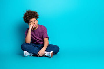 Thoughtful teenage boy sitting cross-legged on blue background wearing casual outfit and looking aside
