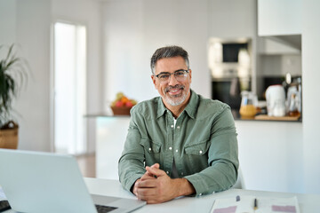 Portrait of happy middle aged senior man wearing glasses sitting with laptop at home table. Smiling older mature handsome man in eyeglasses looking at camera posing with computer sitting in kitchen.
