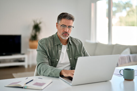 Busy mature middle aged man wearing eyeglasses looking at computer technology sitting at table. Serious older man using laptop hybrid working online, elearning, browsing web, searching online at home.