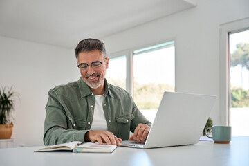 Happy smiling older mature middle aged professional man wearing eyeglasses sitting at living room table using laptop computer hybrid working, elearning, browsing online at casual home.