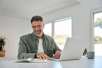 Selbstklebende Fototapeten Happy smiling older mature middle aged professional man wearing eyeglasses sitting at living room table using laptop computer hybrid working, elearning, browsing online at casual home. © insta_photos