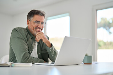 Smiling wearing eyeglasses looking at computer sitting at table. Happy older mature middle aged man using laptop hybrid working online, elearning, browsing web, searching online at home.