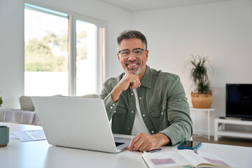 Naklejka na ściany i meble Happy smiling mature middle aged older professional man wearing eyeglasses looking at camera sitting at living room table, using laptop computer working online at casual home. Portrait.