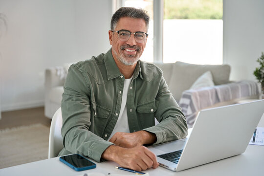 Portrait of happy smiling mature middle aged older professional man wearing glasses looking at camera sitting at living room table, using laptop computer working online at casual home.