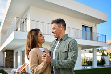 Happy smiling older family couple new property buyers modern villa owners standing outside house, affectionate middle aged man and woman in love hugging outdoors. Vacation home rentals.