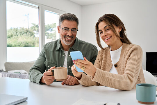 Smiling mature older man and woman holding cellphone browsing internet, texting message on mobile cell phone technology. Middle aged happy couple using smartphone relaxing at living room table at home