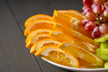 A plate of sliced oranges and grapes on a wooden table. The plate is white and the fruit is...