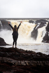 A woman seen from behind stands before the powerful Chaudière Falls, capturing a moment of awe in...