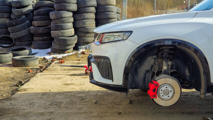 A white car with red rims is parked in front of a pile of tires. The tires are stacked in a heap,...